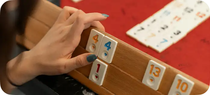 Close-up van de hand van een speler die tegels met cijfers op een houten bord voor het spel Rummikub legt.