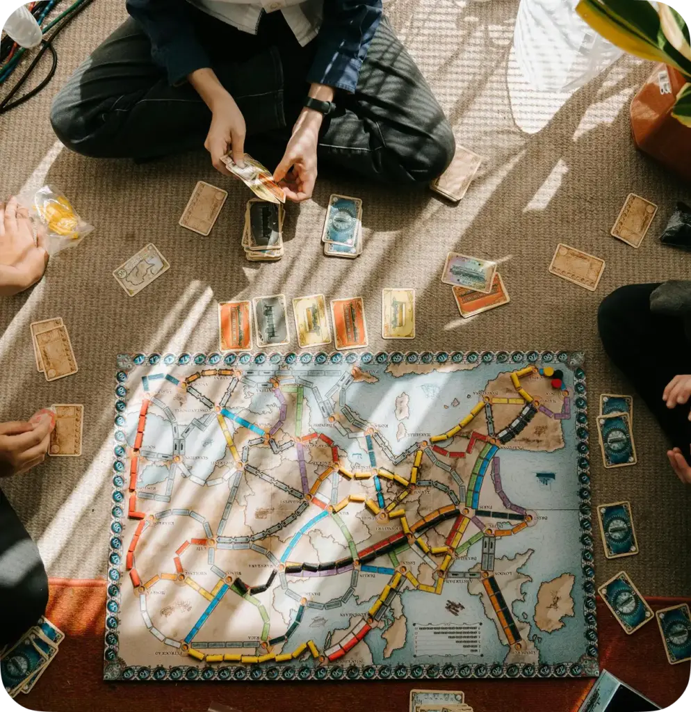 Bovenaanzicht: mensen zitten op een tapijt rond een uitgespreid bordspel Ticket to Ride.